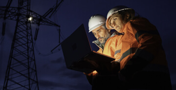Two technicians wearing orange safety gear on a job site at night reviewing a laptop